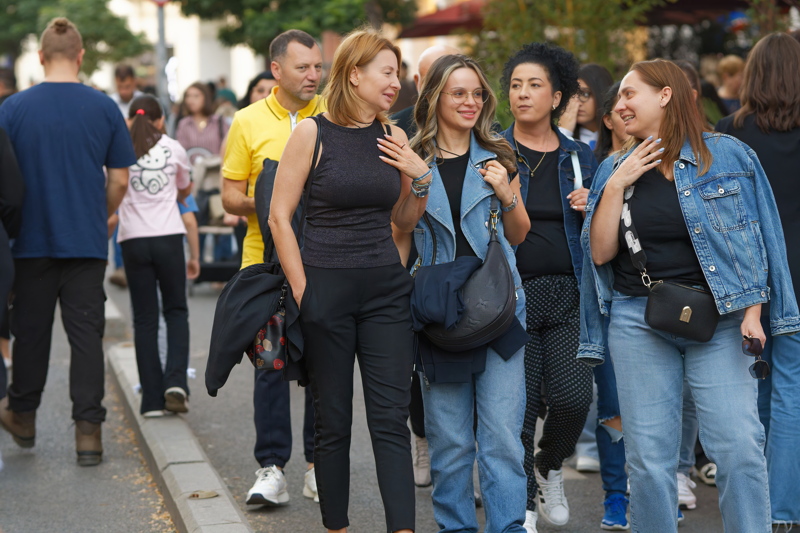 8 - A group of people walking along an outdoor pedestrian street, featuring women wearing casual everyday clothes such as black tops, blue jeans, and denim jackets